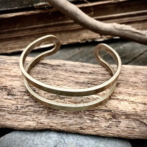 May include: A simple gold-toned metal bracelet with a double band design. The bracelet is lying on a piece of weathered wood.