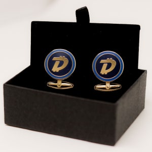 May include: A pair of gold cufflinks with a blue and gold logo of a stylized letter D inside a circle. The cufflinks are in a black velvet box.