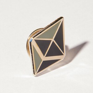 May include: A gold-coloured pin with a black and green geometric design. The pin is shaped like a diamond and has a sharp point at the top.