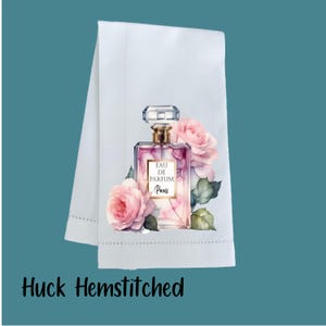 May include: A white huck hemstitched towel featuring a printed graphic of a rectangular glass perfume bottle with a gold cap. The bottle is labeled EAU DE PARFUM Paris and is surrounded by soft pink roses and green leaves on a white fabric.