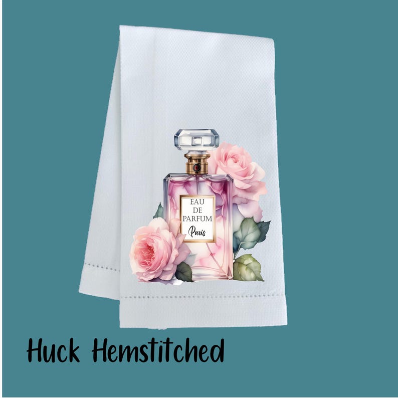 May include: A white huck hemstitched towel featuring a printed graphic of a rectangular glass perfume bottle with a gold cap. The bottle is labeled EAU DE PARFUM Paris and is surrounded by soft pink roses and green leaves on a white fabric.