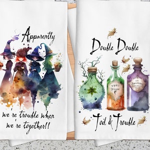May include: Two kitchen towels with a Halloween theme. The first towel features a watercolor illustration of three witches with the text "Apparently we're trouble when we're together!!". The second towel features a watercolor illustration of three potion bottles with the text "Double Double Toil & Trouble".