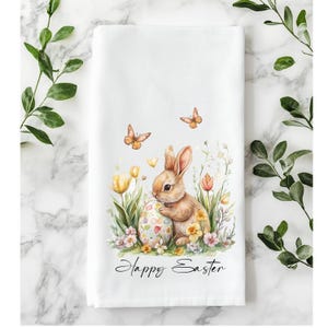 Könnte beinhalten: Weißes Küchentuch zum Thema Ostern mit einer Aquarellillustration eines braunen Hasen, eines verzierten Eies, Schmetterlingen und Blumen. Die Worte "Happy Easter" sind darunter in Schrift geschrieben.