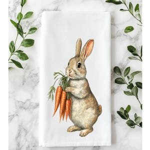 Könnte beinhalten: Weißes rechteckiges Küchentuch mit einer Aquarellillustration eines braunen Kaninchens, das orange Karotten hält. Das Kaninchen hat lange Ohren und steht aufrecht. Grünes Laub befindet sich oben und an den Seiten des Handtuchs.