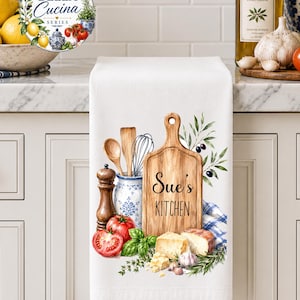 Peut inclure: Un torchon de cuisine blanc orné d'une illustration aquarelle d'une planche à découper en bois avec le texte "Sue's KITCHEN", un pot à ustensiles bleu et blanc avec un fouet et une cuillère, un moulin à poivre, des tomates, du basilic, du fromage et des olives.