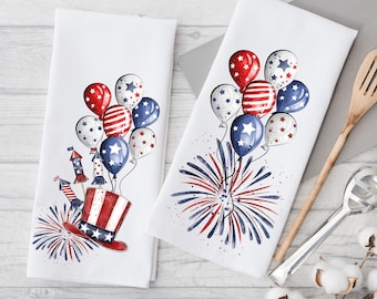 May include: Two white kitchen towels with red, white, and blue watercolor designs. One towel features a red, white, and blue top hat with fireworks and balloons. The other towel features a cluster of red, white, and blue balloons with fireworks.