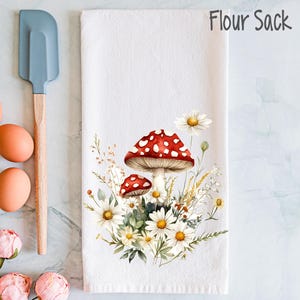 Op de afbeelding: Witte keukendoek van meelzak met een rode en witte paddenstoel en witte madeliefjes erop gedrukt. De tekst "Flour Sack" is gedrukt in de rechterbovenhoek.
