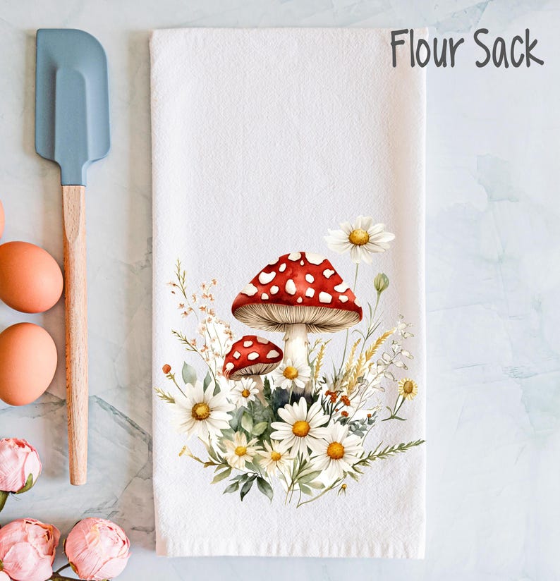 Op de afbeelding: Witte keukendoek van meelzak met een rode en witte paddenstoel en witte madeliefjes erop gedrukt. De tekst "Flour Sack" is gedrukt in de rechterbovenhoek.
