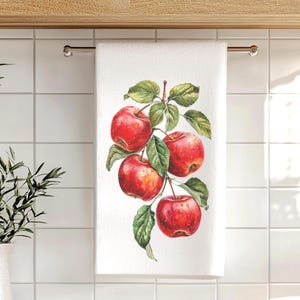 May include: A white kitchen towel featuring a detailed botanical illustration of four bright red apples hanging from a brown branch with several green leaves. The apples appear to have small water droplets on their skins, adding a realistic texture to the printed design on the fabric.