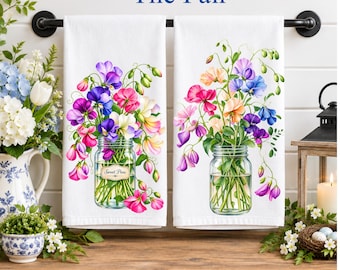 Op de afbeelding: Twee witte keukendoeken hangen aan een zwarte stang, elk met een afbeelding van een glazen pot gevuld met kleurrijke pronkerwten. De linkerdoek heeft een klein label op de pot met de tekst Sweet Peas.