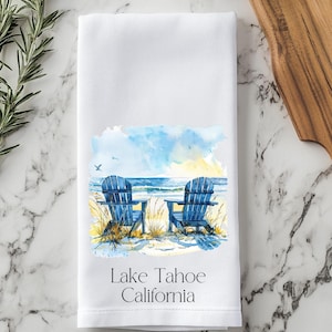 May include: White kitchen towel with a watercolor illustration of two blue Adirondack chairs on a sandy beach with a lake or ocean in the background. The text "Lake Tahoe California" is printed below the image.