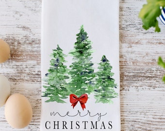 Peut inclure: Une serviette de cuisine blanche avec une illustration aquarelle de trois sapins de Noël verts avec un nœud rouge au centre. Le texte "merry christmas" est écrit sous les arbres.