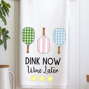Puede incluir: Toalla de cocina blanca con raquetas de paddleball a cuadros verdes, rosas y azules y el texto "DINK NOW Wine Later" con tres pelotas de paddle amarillas debajo.
