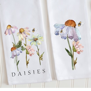 Op de afbeelding: Twee witte theedoeken met aquarel illustraties van madeliefjes en bijen. De tekst "DAISIES" is op een theedoek gedrukt.