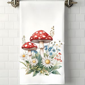 Op de afbeelding: Een witte keukendoek met een aquarel-illustratie van rode en witte paddenstoelen en blauwe en witte bloemen.