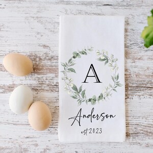 Peut inclure: Une serviette de cuisine blanche avec une couronne de fleurs vertes et la lettre A au centre. Le texte "Anderson est 2023" est sous la couronne.
