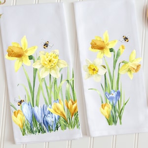 May include: Two white tea towels with a watercolor design of yellow daffodils, blue crocuses, and green leaves.  There are also two small bees on each towel.