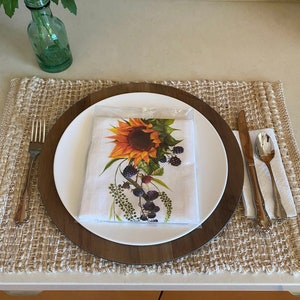May include: A white dinner plate with a brown wooden charger sits on a woven placemat. The plate features a sunflower and blackberry design. A fork, knife, and spoon are placed to the right of the plate.