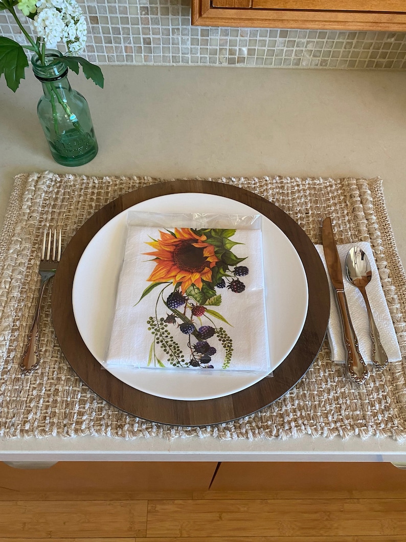 May include: A white dinner plate with a brown wooden charger sits on a woven placemat. The plate features a sunflower and blackberry design. A fork, knife, and spoon are placed to the right of the plate.