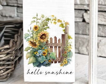 May include: A white kitchen towel with a watercolor illustration of sunflowers, a wooden fence, and green leaves. The text "hello sunshine" is printed in black cursive font at the bottom of the towel.