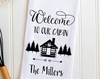 May include: White kitchen towel with black text and a cabin illustration. The text reads "Welcome to our cabin" and "The Millers".