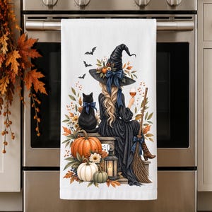 May include: A white kitchen towel featuring a colorful illustration of a witch with long blonde hair wearing a black hat and dress, sitting with a black cat, pumpkins, a lantern, a broom, and bats, while holding a glass of orange liquid.