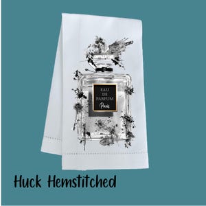 May include: White Huck hemstitched towel featuring a perfume bottle design. The perfume bottle is clear with a black and white abstract design and the words "EAU DE PARFUM Paris".