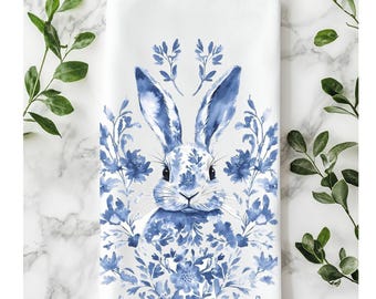 May include: White rectangular kitchen towel with a blue watercolour illustration of a rabbit surrounded by blue floral elements. The rabbit has long ears and a detailed face. The towel is decorated with a floral border and is surrounded by green leaves.