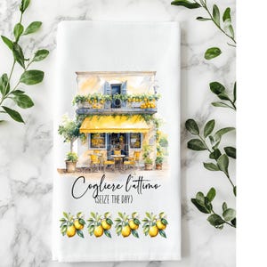 May include: White kitchen towel with a watercolor illustration of a cafe with a yellow awning and lemons. The text "Cogliere l'attimo (Seize the Day)" is written in script. A border of lemons and leaves decorates the bottom.