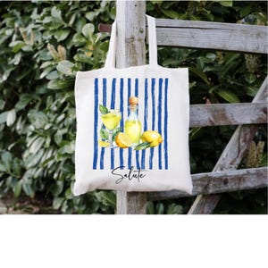 May include: White tote bag with blue vertical stripes and a watercolor illustration of lemons, a bottle, and a glass of yellow liquid. The word "Salute" is written in cursive script below the illustration.