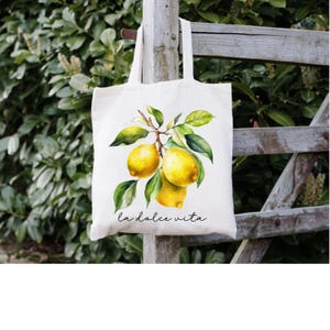 May include: Cream-colored tote bag featuring a watercolor illustration of lemons on a branch with green leaves. The text "la dolce vita" is written in a script font below the lemons. The bag has two handles and is hanging on a wooden fence.