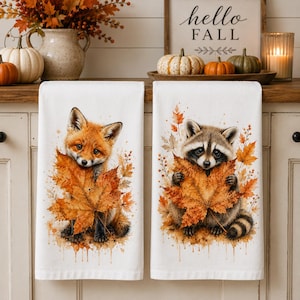 May include: Two white kitchen towels featuring watercolor-style illustrations. The left towel displays a red fox holding a large orange maple leaf. The right towel shows a raccoon holding a similar orange maple leaf, surrounded by autumn foliage.