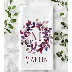 May include: White kitchen towel featuring a watercolor wreath of purple and burgundy leaves around a large letter "M". The name "Martin" is printed in a matching burgundy font below the wreath. Decorative greenery surrounds the towel.
