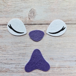 May include: Felt craft pieces arranged to resemble a face. The set includes two white teardrop-shaped pieces with black curved lines, a small purple oval, and a larger purple shape resembling a nose.