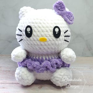 May include: White crocheted Hello Kitty plush toy with purple bow and skirt. The toy has black eyes and a yellow nose.