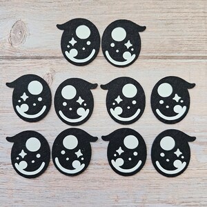 May include: Ten black felt eye shapes with white accents, arranged on a light wood surface. Each eye has a white circle, starbursts, and a curved white smile. The shapes are symmetrical and suitable for crafting or embellishment.