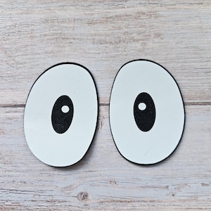 May include: Two white googly eyes with black pupils and white highlights. The eyes are on a light brown wooden background.