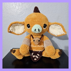 May include: A crocheted plush toy in the shape of a brown and white creature with a green nose and large eyes. The toy is wearing a brown and tan necklace with a small crocheted brown and white creature hanging from it. The toy is sitting on a dark surface. "Brunsheka Designs" is written on the toy's necklace.