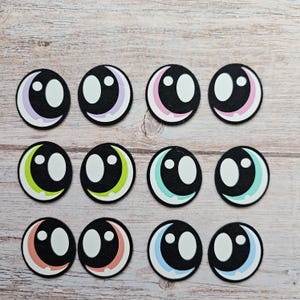 May include: Twelve colorful felt eyes with black pupils and white sclera. The eyes have a crescent moon shape around the pupil in various colors including pink, purple, green, blue, and orange.