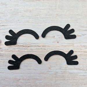 May include: Four black felt eyelashes with a curved shape and three points on the outer edge. The eyelashes are arranged in two pairs on a light wood background.