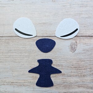 May include: Arrangement of felt shapes for a craft project. Includes two white, teardrop-shaped pieces with black curved lines, a navy blue oval, and a navy blue figure resembling a bow tie. The pieces are arranged on a light brown wooden surface.