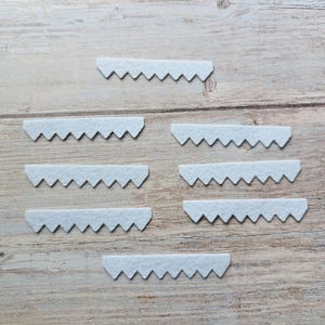 May include: Seven light blue felt sawtooth picture hangers. Each hanger has a series of triangular teeth along one edge. The hangers are arranged on a light brown wooden surface.