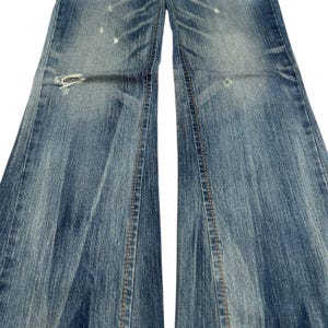 May include: Close-up of distressed blue denim jeans. The jeans have a faded wash, visible wear, and a ripped section near the knee. The seams are stitched with a contrasting tan thread.