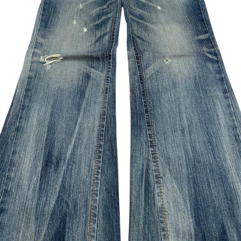 May include: Close-up of distressed blue denim jeans. The jeans have a faded wash, visible wear, and a ripped section near the knee. The seams are stitched with a contrasting tan thread.