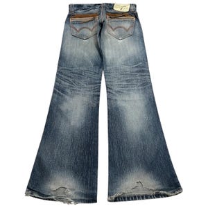 May include: Faded blue denim jeans with a wide leg design. The jeans feature brown suede-like accents on the back pockets and a distressed look at the hem. A small tag is visible at the back of the waistband.
