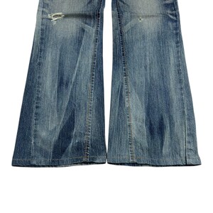 May include: Close-up of light blue denim jeans with a flared leg design. The jeans have distressed details, including a ripped section near the knee. The denim has a faded appearance, with visible stitching and a classic five-pocket style.