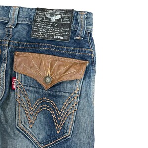 May include: Close-up of blue denim jeans with a brown leather pocket and decorative stitching. The jeans have a label that reads "MADE IN JAPAN" and "EDWIN SU". The back pocket features a unique design with a button closure.