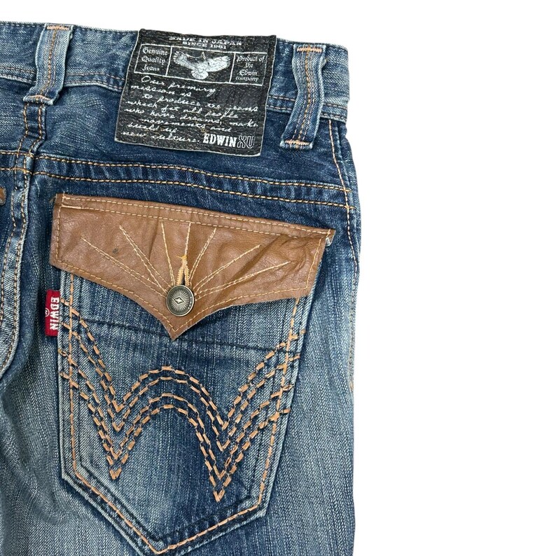 May include: Close-up of blue denim jeans with a brown leather pocket and decorative stitching. The jeans have a label that reads "MADE IN JAPAN" and "EDWIN SU". The back pocket features a unique design with a button closure.