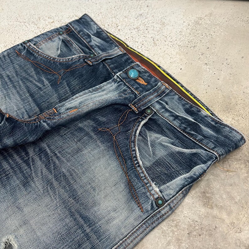 May include: Close-up of blue denim jeans with orange stitching. The jeans have a button closure and a distressed look. The waistband features a yellow and brown stripe.