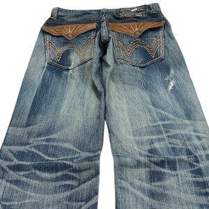 May include: Faded blue denim jeans with distressed detailing. Features brown leather-look back pockets with decorative stitching and a branded patch at the waistband. The jeans have a relaxed fit and a unique wash pattern.